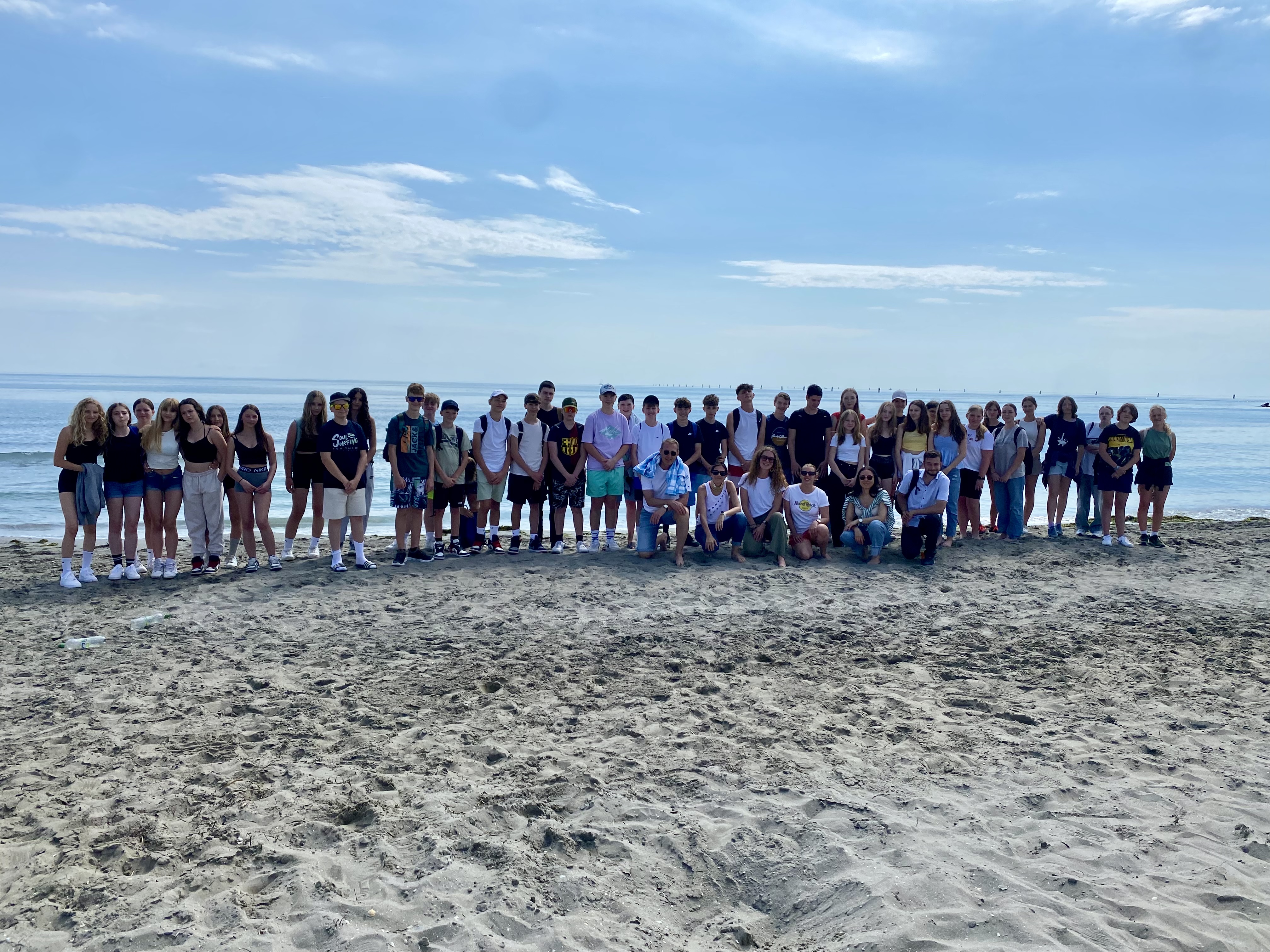 Eine Gruppe Schüler und Schülerinnen steht mit ihren Lehrer:innen am Sandstrand von Grado bei schönem Wetter 